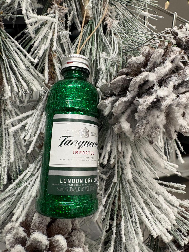 Puede incluir: Adorno navide&ntilde;o en forma de botella de ginebra en miniatura, cubierta de purpurina verde. La etiqueta dice "Tanqueray Imported London Dry Gin", con un volumen de 50 ml. El adorno cuelga de una cuerda, sobre un fondo de pino helado y una pi&ntilde;a.