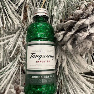 Puede incluir: Una botella en miniatura de ginebra Tanqueray London Dry cubierta de purpurina verde, un adorno festivo. La botella tiene una tapa plateada y una etiqueta blanca con texto negro. El fondo presenta agujas de pino escarchadas y una pi&ntilde;a.