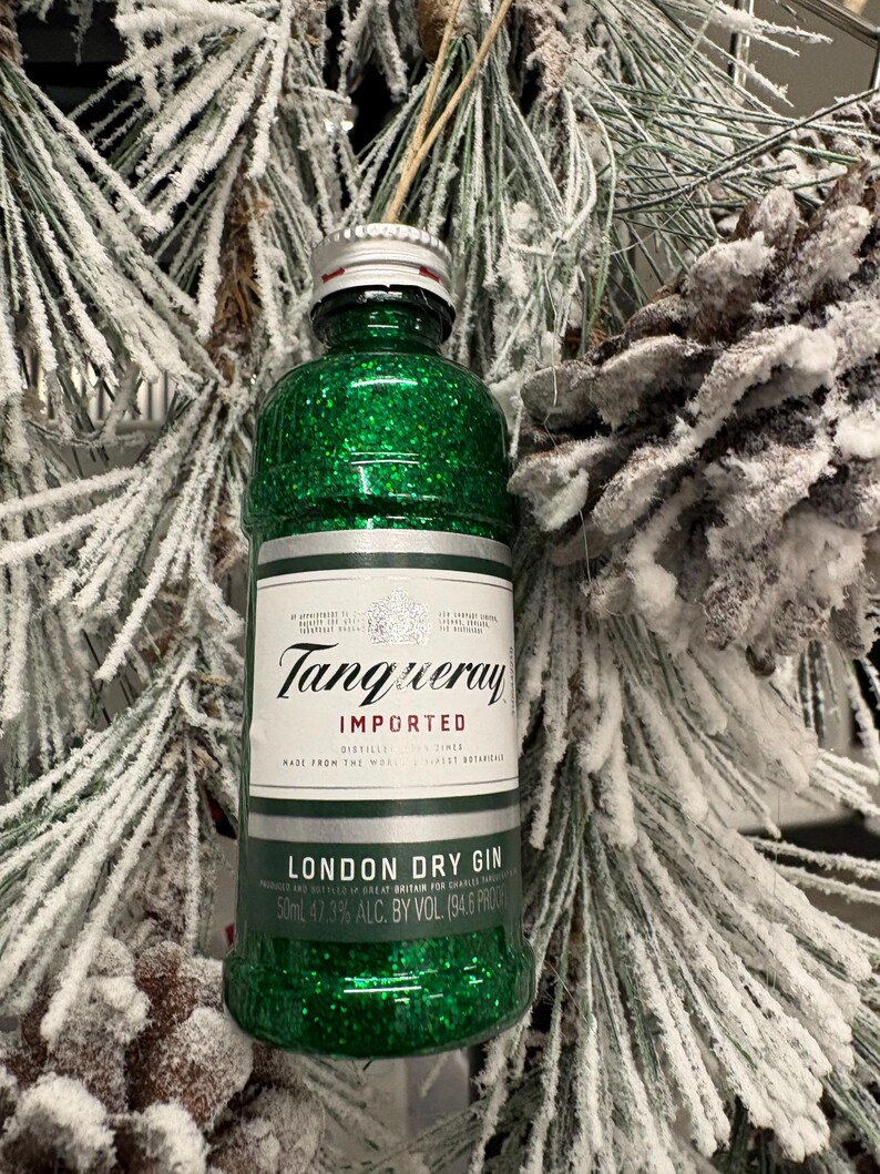 Puede incluir: Una botella en miniatura de ginebra Tanqueray London Dry cubierta de purpurina verde, un adorno festivo. La botella tiene una tapa plateada y una etiqueta blanca con texto negro. El fondo presenta agujas de pino escarchadas y una pi&ntilde;a.