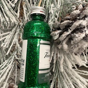Puede incluir: Adorno navide&ntilde;o de botella verde brillante con tap&oacute;n plateado y etiqueta "Tanqueray London Dry Gin". El adorno cuelga de una cuerda natural, sobre un fondo de ramas de pino escarchadas.