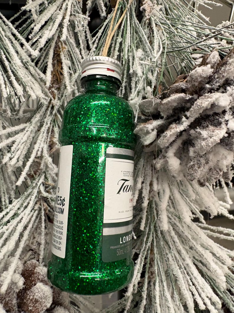 Puede incluir: Adorno navide&ntilde;o de botella verde brillante con tap&oacute;n plateado y etiqueta "Tanqueray London Dry Gin". El adorno cuelga de una cuerda natural, sobre un fondo de ramas de pino escarchadas.