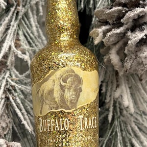 Puede incluir: Adorno de botella en miniatura cubierto de purpurina con una etiqueta que dice "Buffalo Trace Kentucky Straight Bourbon Whiskey". La botella tiene una tapa verde y un acabado de purpurina dorada. El adorno cuelga de una cuerda.