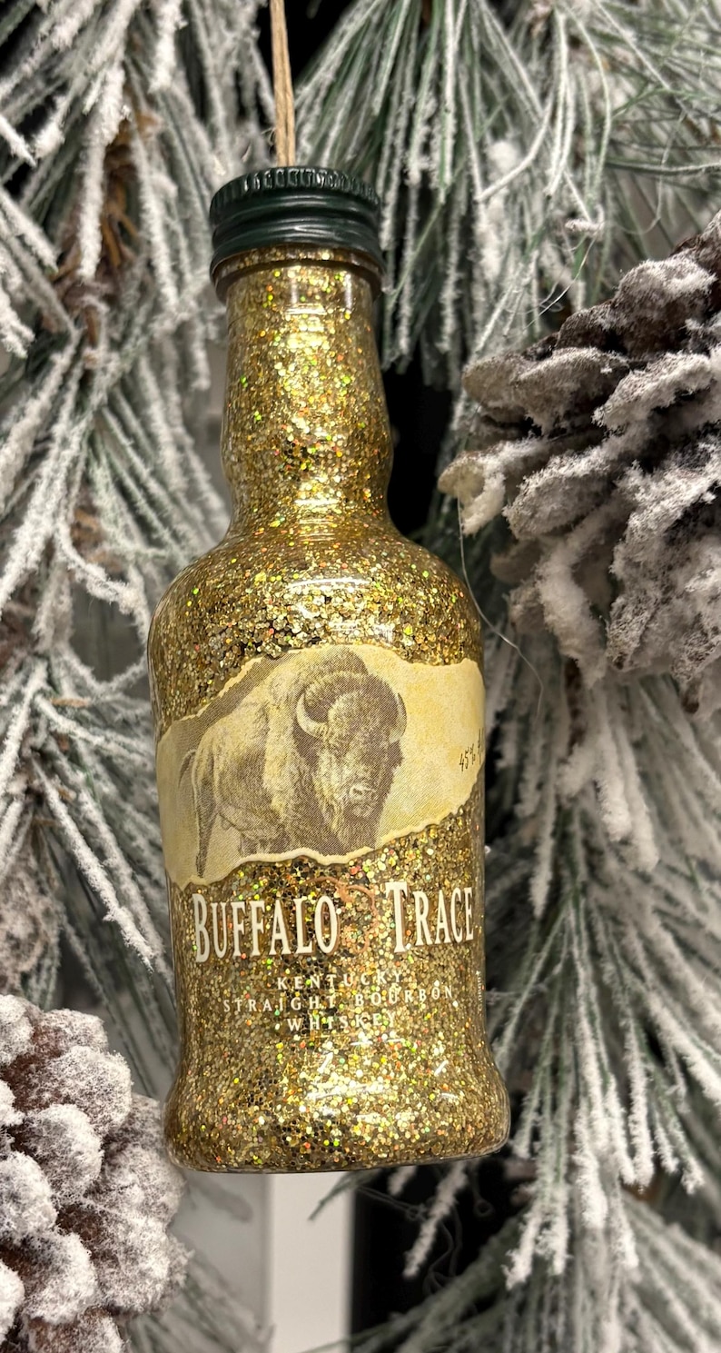 Puede incluir: Adorno de botella en miniatura cubierto de purpurina con una etiqueta que dice "Buffalo Trace Kentucky Straight Bourbon Whiskey". La botella tiene una tapa verde y un acabado de purpurina dorada. El adorno cuelga de una cuerda.