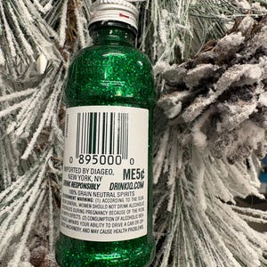 Puede incluir: Adorno navide&ntilde;o en forma de botella verde brillante con tapa plateada y etiqueta. La etiqueta incluye un c&oacute;digo de barras y el texto "IMPORTED BY DIAGEO, NEW YORK, NY". El adorno cuelga de una rama de pino nevada.