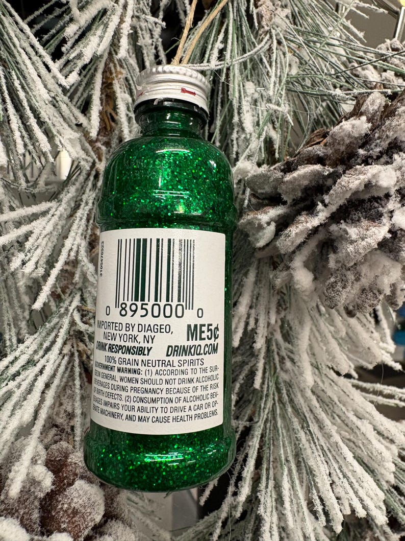 Puede incluir: Adorno navide&ntilde;o en forma de botella verde brillante con tapa plateada y etiqueta. La etiqueta incluye un c&oacute;digo de barras y el texto "IMPORTED BY DIAGEO, NEW YORK, NY". El adorno cuelga de una rama de pino nevada.