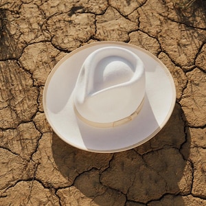 May include: A white fedora hat with a wide brim and a cream coloured band. The hat is sitting on a cracked, dry dirt surface.