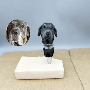 Może przedstawiać: Czarny korek do butelki z głową psa, srebrną podstawą i czarną gumową uszczelką. Głowa psa jest szczegółowa z realistycznymi cechami. Po lewej stronie znajduje się okrągły obraz twarzy psa.