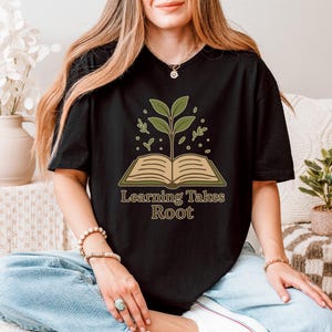 May include: Black t-shirt featuring an open book with a plant growing from it, and the text "Learning Takes Root" in a brown font. The shirt is worn by a person in blue jeans.