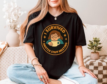 May include: Black t-shirt with a circular graphic that reads "I Survived Another Staff Meeting." The graphic features a cartoon person with glasses looking sad and holding a yellow mug. The shirt is worn by a person.