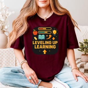 May include: A maroon t-shirt featuring the text "Leveling Up Learning" in a graphic design. The design includes an apple, a book, a lightbulb, and a progress bar, with an upward arrow at the bottom.
