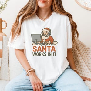 Peut inclure: T-shirt blanc avec un dessin de Père Noël de dessin animé portant un casque et travaillant sur un ordinateur. Le texte "SANTA WORKS IN IT" est imprimé en dessous en rouge et vert. Le t-shirt est de conception décontractée et festive.