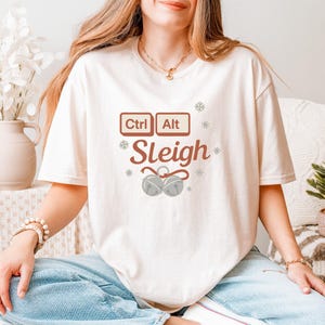 Peut inclure: T-shirt crème avec un motif festif. Le graphisme comprend les mots "Ctrl Alt Sleigh" avec des touches de clavier et des flocons de neige. En dessous, deux cloches argentées. Un t-shirt de vacances amusant.