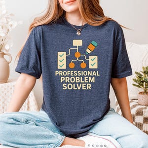 May include: Navy blue t-shirt with the text "PROFESSIONAL PROBLEM SOLVER" and a diagram graphic. The graphic includes a pencil, check marks, and orange circles. The shirt is made of a soft material.