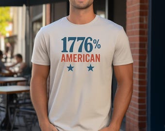 May include: A light grey t-shirt with the text "1776% AMERICAN" in blue and red, with two blue stars below. The shirt is short-sleeved and appears to be made of a soft material.