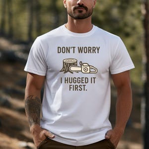 May include: White t-shirt with the text "DON'T WORRY I HUGGED IT FIRST." and a graphic of a tree stump and chainsaw. The design is in brown and beige tones. The shirt is a crew neck style.