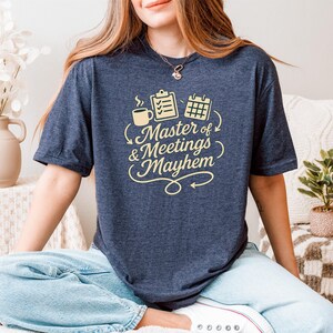 May include: A navy blue t-shirt with the text "Master of Meetings & Mayhem" in a cream-coloured script. The design includes a coffee cup, clipboard, and calendar. The shirt is worn by a person sitting cross-legged.