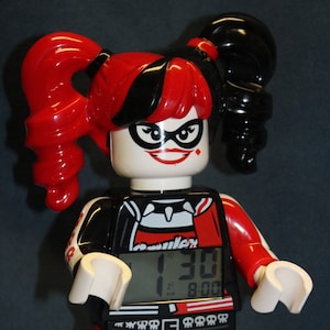 May include: A red and black Lego clock featuring Harley Quinn with a digital display showing the time 1:30. The clock has a white face with black numbers and a black background. The clock is set to 8:00.