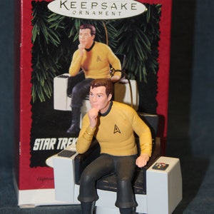 May include: A red box with a Star Trek ornament inside. The ornament is a figurine of Captain Kirk sitting in the captain's chair. The box has the words "Keepsake Ornament" and "Star Trek" on it.