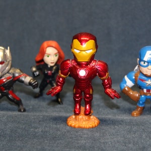 Puede incluir: Cuatro figuras de superhéroes: Ant-Man en negro y rojo, Black Widow en negro, Iron Man en rojo y dorado, y Capitán América en azul y marrón. Las figuras están estilizadas con rasgos exagerados.
