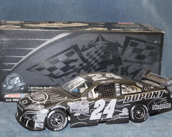 May include: A 1:24 scale die-cast stock car model featuring a black finish with white flame graphics and the number 24. The car displays sponsor logos including DuPont, Nicorette, and Pepsi, and is presented in front of its original black cardboard box.