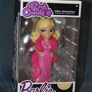 May include: A pink and white vinyl collectible figure of Barbie in a pink dress and a pink fur stole. The figure is in a clear plastic box with the words "Rock Candy" and "Barbie" on the front. The box also has the year "1977" and the words "Vinyl Collectible" on the front.