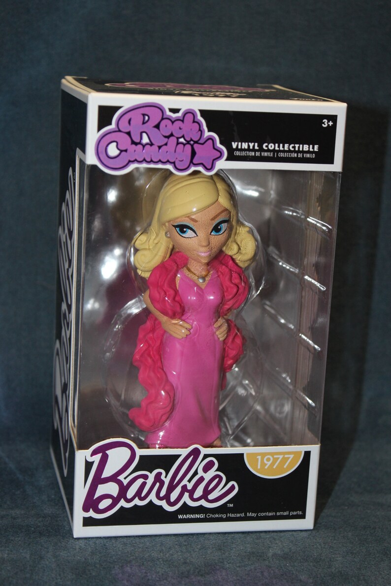 May include: A pink and white vinyl collectible figure of Barbie in a pink dress and a pink fur stole. The figure is in a clear plastic box with the words "Rock Candy" and "Barbie" on the front. The box also has the year "1977" and the words "Vinyl Collectible" on the front.