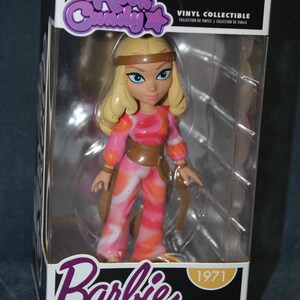 May include: A vinyl collectible figure of Barbie in a pink and orange patterned outfit with a brown belt. The figure is in a clear plastic box with a white background. The box has the words "Rock Candy" and "Barbie" on it, along with the year "1971".