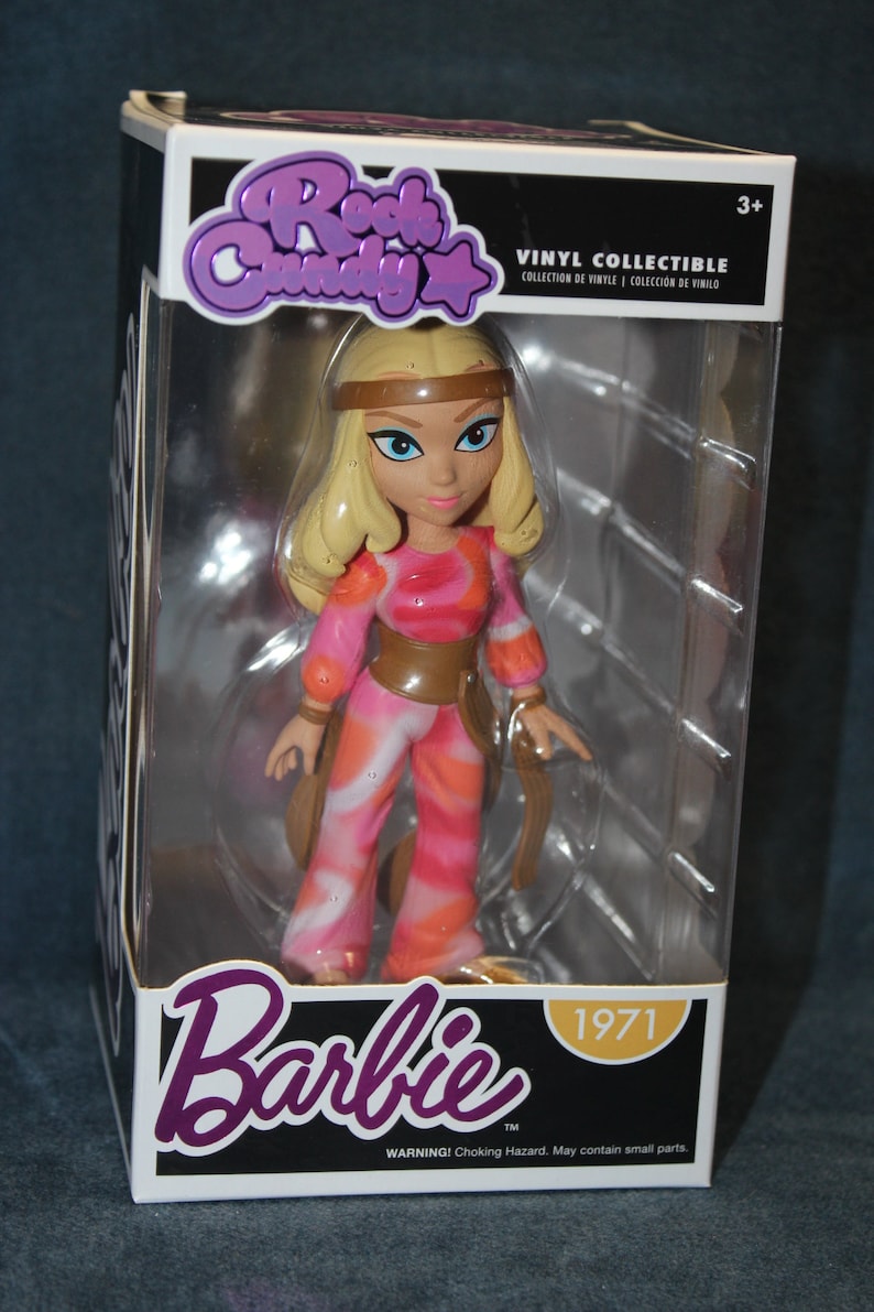 May include: A vinyl collectible figure of Barbie in a pink and orange patterned outfit with a brown belt. The figure is in a clear plastic box with a white background. The box has the words "Rock Candy" and "Barbie" on it, along with the year "1971".