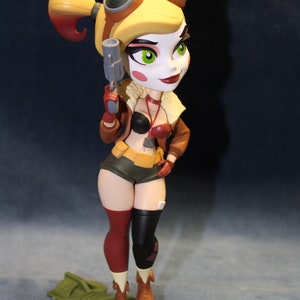 May include: A small, stylized figurine of Harley Quinn in a pilot's outfit, holding a gun and standing on a bomb-shaped base. She has red and black leggings, a brown leather jacket, and a yellow and red hair style.