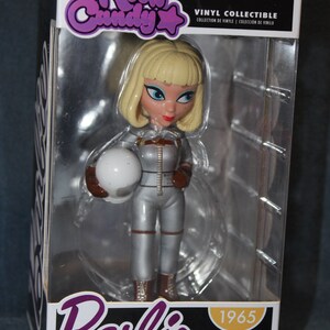 May include: A vinyl collectible figure of a Barbie doll dressed in a silver space suit holding a white helmet. The figure is in a clear plastic box with a black and white background. The box has the words "Rock Candy" and "Barbie" on it. The box also has the year "1965" on it.