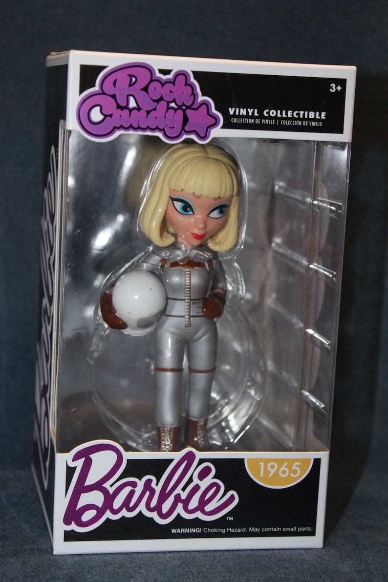 May include: A vinyl collectible figure of a Barbie doll dressed in a silver space suit holding a white helmet. The figure is in a clear plastic box with a black and white background. The box has the words "Rock Candy" and "Barbie" on it. The box also has the year "1965" on it.