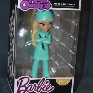May include: A vinyl collectible figure of Barbie dressed as a doctor. She is wearing a teal scrub top and pants, a white hat, and a stethoscope. The figure is in a clear plastic box with a white background. The box has the words "Rock Candy" and "Barbie" on it, along with the year "1973".