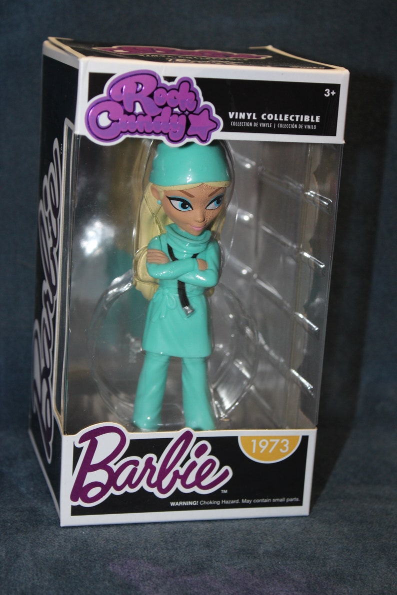 May include: A vinyl collectible figure of Barbie dressed as a doctor. She is wearing a teal scrub top and pants, a white hat, and a stethoscope. The figure is in a clear plastic box with a white background. The box has the words "Rock Candy" and "Barbie" on it, along with the year "1973".