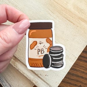 Può includere: Un adesivo raffigurante un barattolo di burro di arachidi con le lettere "PB" e due arachidi. Accanto al barattolo c'è una pila di biscotti sandwich al cioccolato. Il barattolo è marrone con un coperchio marrone scuro.