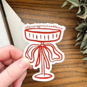 May include: Red vinyl sticker with a hand-drawn illustration of a champagne bottle with a bow and the text "Champagne problems".