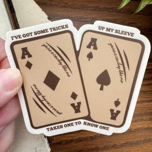May include: A sticker showing two playing cards, each with an Ace. The cards are tan with brown borders and the text "I've got some tricks" and "Up my sleeve." The phrase "you're a cowboy like me" is also printed on the cards.