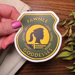 Może przedstawiać: Naklejka w kształcie tarczy z napisem "PAWNEE GODDESSES". Tarcza jest zielona ze złotą obwódką i złotym kołem pośrodku. Wewnątrz koła znajduje się czarna sylwetka kobiety.