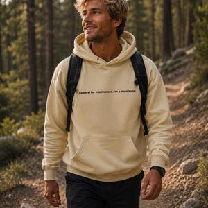 May include: A cream-colored hooded sweatshirt featuring a front kangaroo pocket and black text that reads, "Apparel for manifestors. I'm a manifestor." The long-sleeved pullover is worn with black shorts and a black backpack with shoulder straps.