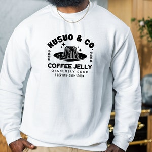 May include: White sweatshirt with a black graphic that reads "Kusuo & Co. Coffee Jelly." The design includes an illustration of coffee jelly on a plate, with Japanese characters. The text "Obscenely Good" is below the coffee jelly illustration.