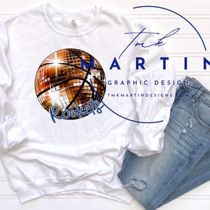 May include: White sweatshirt with a gold disco ball basketball graphic and the text "Rockets" in blue. The sweatshirt is laying on a white wooden surface with a pair of blue jeans with distressed details.