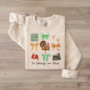 Könnte beinhalten: Ein beiger Pullover mit einem Design aus einem Truthahn, Schleifen und Jagdausrüstung. Der Text "In Spring, we Strut" ist auf dem Pullover aufgedruckt.