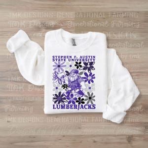 May include: White long-sleeve sweatshirt with a purple, black, and gray graphic design featuring a cartoon lumberjack holding a saw. The graphic also includes the text "Stephen F. Austin State University" and "Lumberjacks".