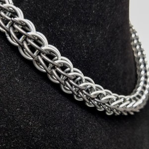 Puede incluir: Un collar de cadena de plata hecho de anillos de metal entrelazados. La cadena está sobre un fondo de terciopelo negro.