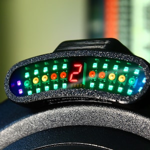 Op de afbeelding: Een zwart elektronisch apparaat met een digitale display die het nummer 2 in het rood weergeeft. De display is omgeven door een reeks groene, gele en rode LED-lampjes. Het apparaat heeft een getextureerd oppervlak.