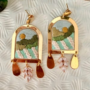 May include: A pair of gold-toned arch-shaped earrings featuring embroidered landscape scenes with a yellow sun, green hills, and white textured fields. Each earring includes three dangling charms: two smooth gold teardrops and a central crystal leaf cluster.