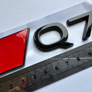 May include: Black Q7 emblem with a red diamond-shaped accent. The emblem is glossy. A ruler is placed next to the emblem for scale. The Q7 emblem is likely for a car. The ruler shows measurements in centimetres.