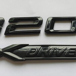 May include: A set of black car emblems, comprising the numbers "320d" and an "X DRIVE" badge. The emblems have a glossy finish and are likely made of plastic or metal. The "X" in "X DRIVE" is a prominent, stylized design.
