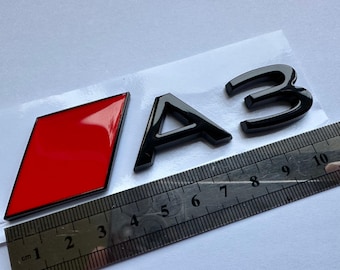 Könnte beinhalten: Schwarzes "A3"-Emblem mit einem roten rautenförmigen Logo. Das Emblem ist glänzend. Ein Lineal mit Zentimetermessungen befindet sich im Vordergrund.