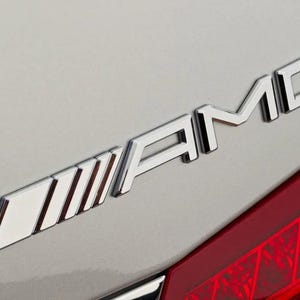 May include: A silver AMG emblem with a series of parallel lines on the left side of the logo. The emblem is attached to the back of a silver car.