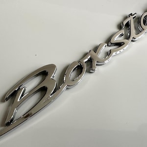 May include: A chrome-finished metal emblem featuring the word Boxster written in a stylised, cursive script font. The letters are connected and have a reflective, polished surface, designed to be mounted as a decorative badge on a vehicle.
